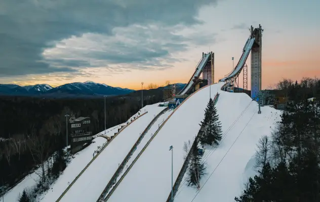 An aerial view of two ski jumps on top of a hill, with a gentle sunrise in the background.