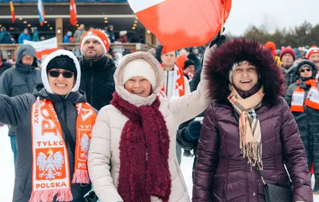 Group of three women stand next to each other bundled heavily in winter clothing at outdoor event with Polish flag and accessories