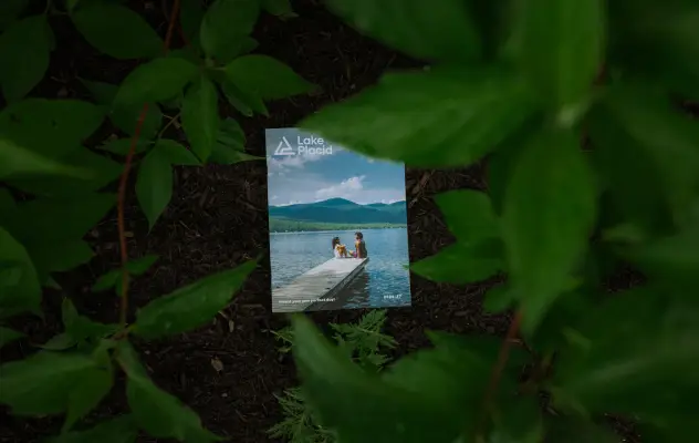 Copy of Lake Placid 2026 Traveler Guide sits on dirt and mulch under green leaves