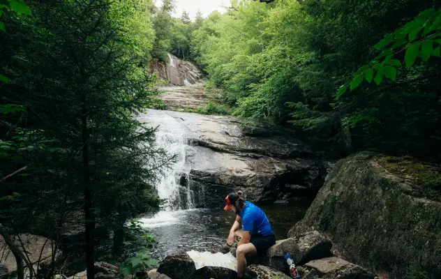 A man reads a map at the base of a waterfall in the woods. 