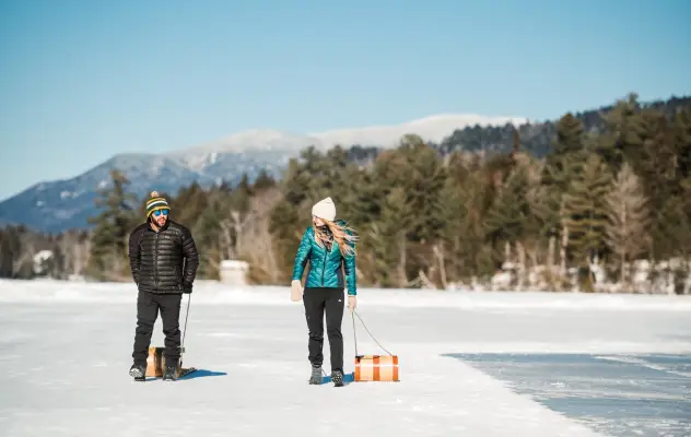 Couple pulls toboggans behind them as they walk across a frozen lake with snow-capped mountains in the background