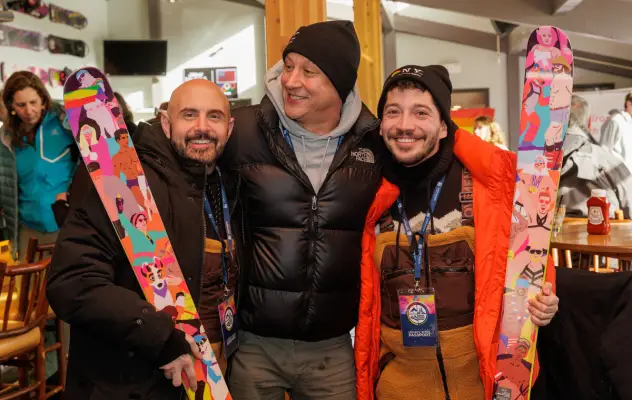 Three men stand together with their arms around one another; the two men on the end are looking at the camera, each holding a colorful ski