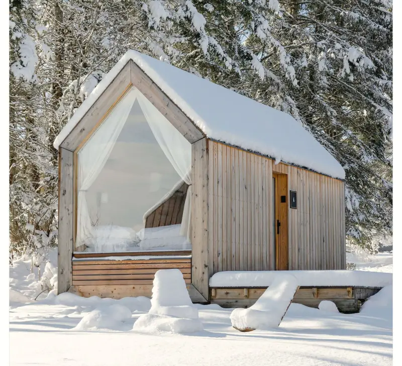 A minimum cabin blanketed in snow surrounded by tall pines