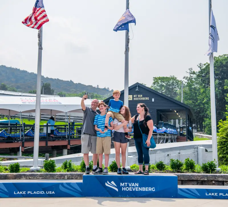 Mother, father, daughter, and older son stand with youngest son on their shoulders taking a selfie on 1st place podium at Mt Van Hoevenberg sliding complex