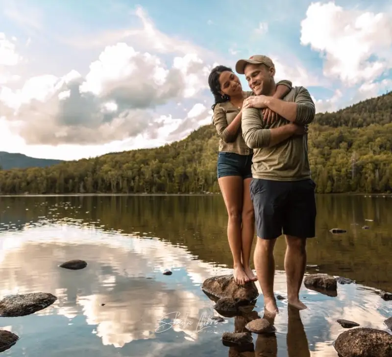 couple-embracing-at-waters-edge-with-mountains-and-blue-skies-in-the-background