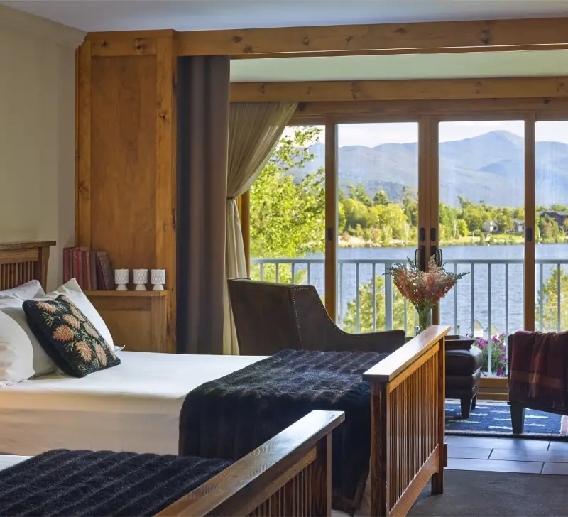 Beautifully appointed bedroom space with adirondack decor like snowshoes adorn the walls and the balcony windows open showing Mirror Lake and the majestic mountainscape background