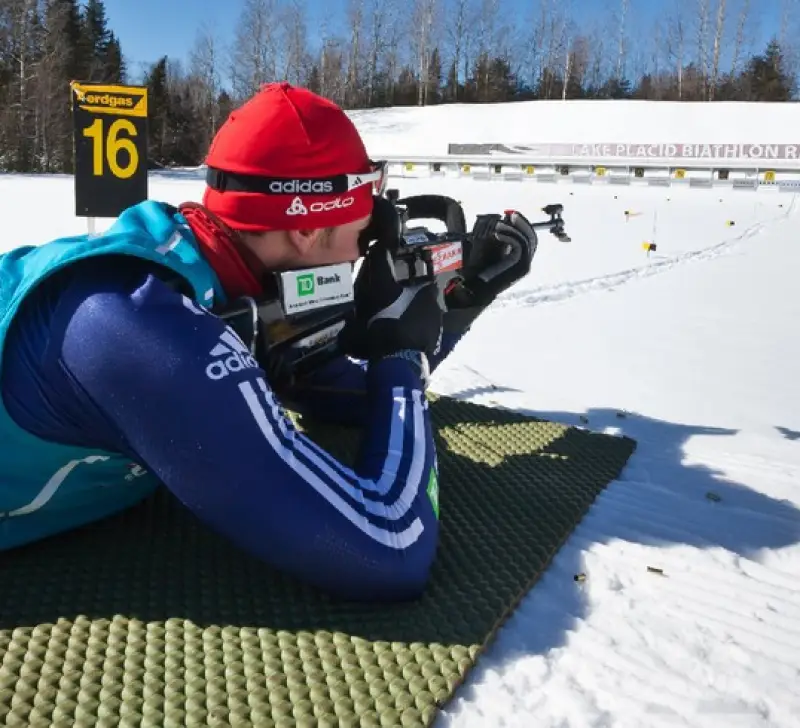 A biathlete takes aim with a rifle while lying down on a pad on snow.
