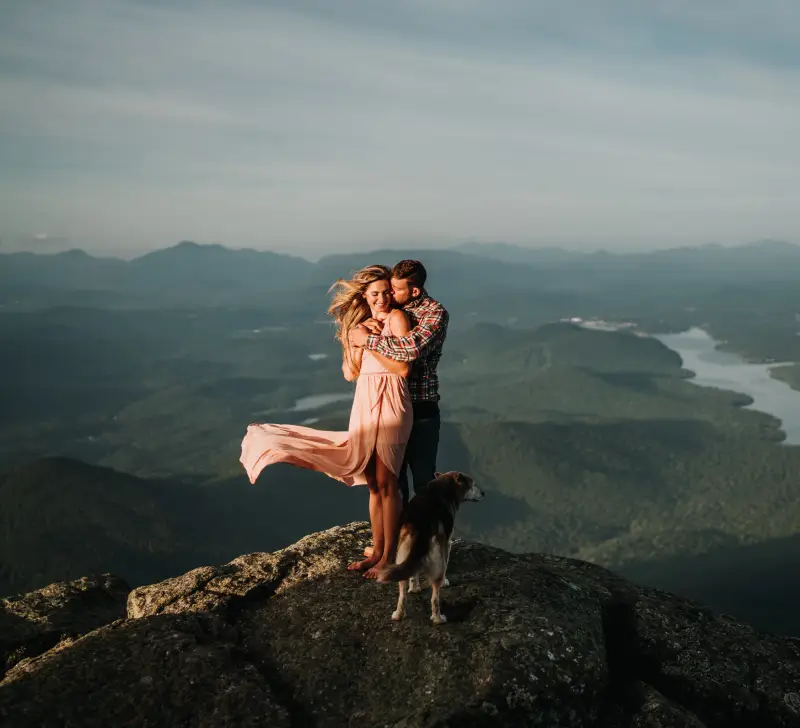 A couple embraces on a rocky ledge while her dress and hair flow in the breeze with Lake Placid in the back drop