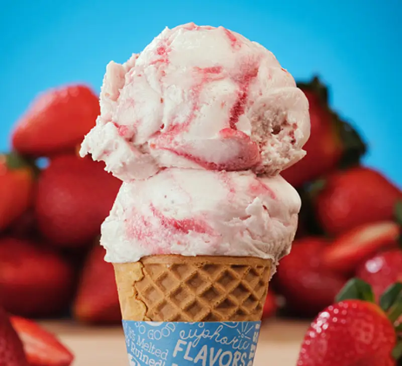 Double scoop of creamy strawberry ice cream in a waffle cone with blue background and ringed with red juicy strawberries