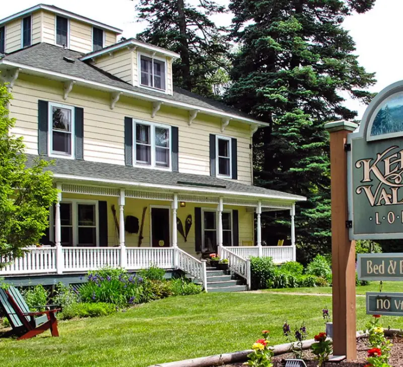 The front of Keene Valley Lodge, a pale yellow house with white trim and a large front porch with the sign seen out front