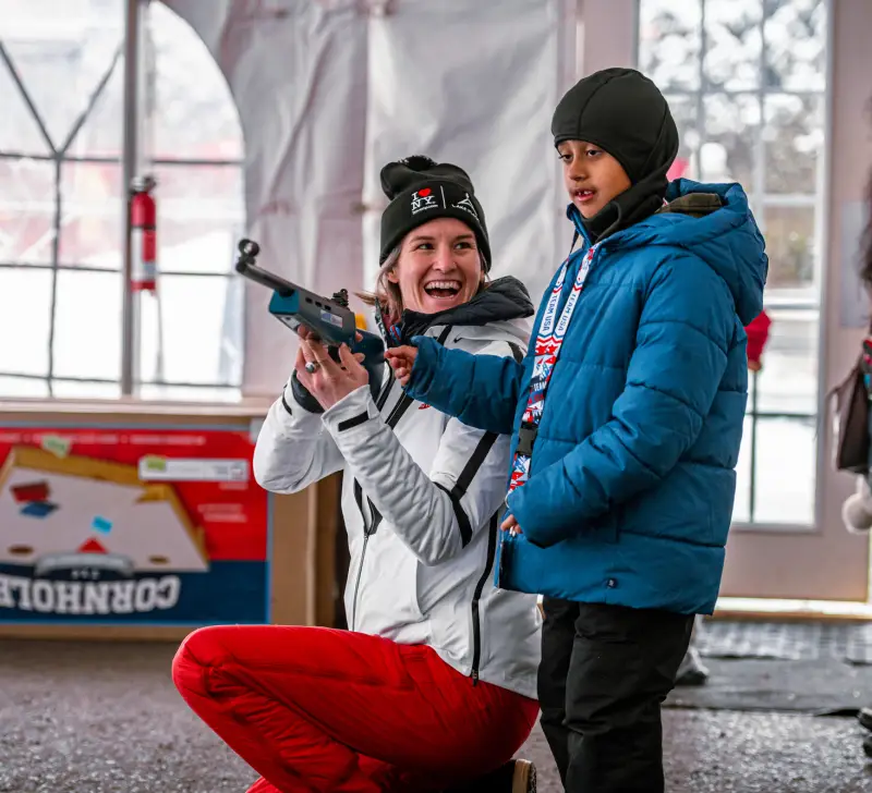 A female biathlete shows her rifle to a child at a biathlon event.