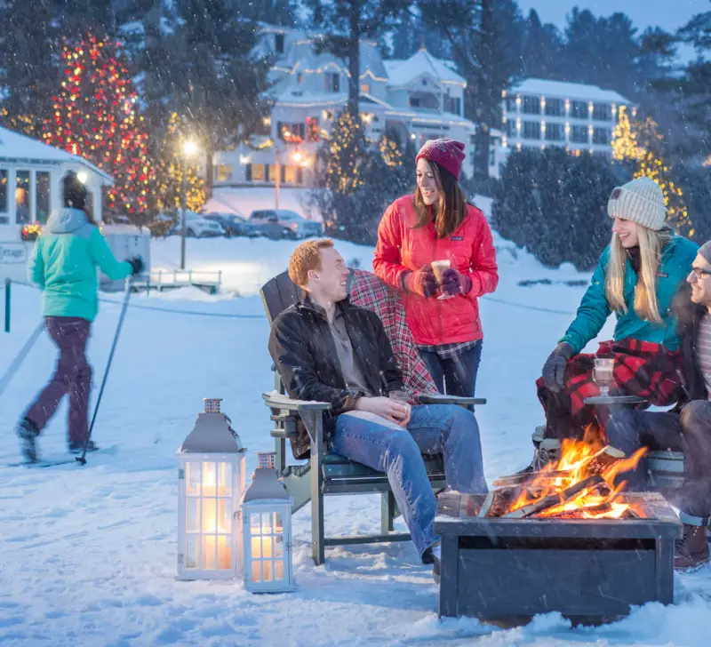 Happy people sit and chat around a fire pit with the glow of christmas lights dot the background and a skiier slides by.