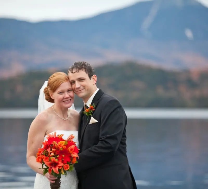 A happy couple smiles into the camera with the Mirror Lake and fall foliage covered mountains in the background