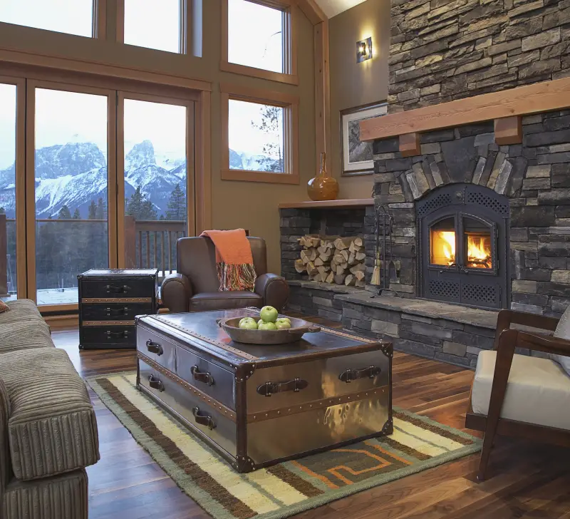 The main living space of a large home with two stories of windows, a stone fireplace with built in wood storage and ample lush seating.