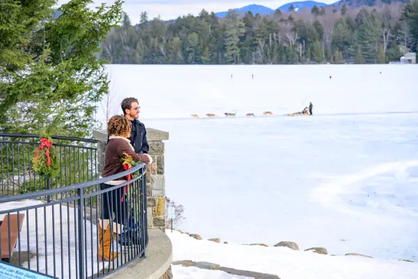 A man and a woman stand on an outdoor balcony, while a dog sled races across the frozen lake in the background.