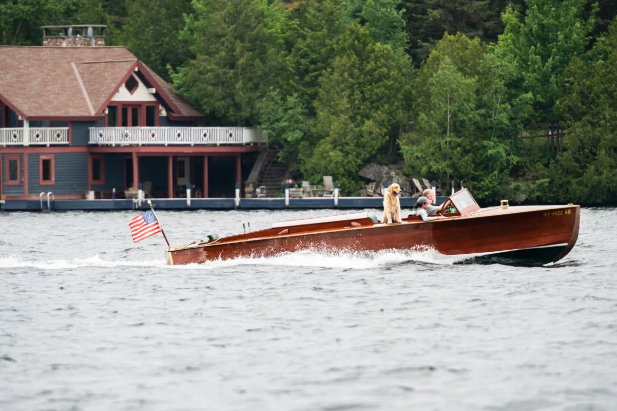 A vintage wooden speedboat carrying two humans and a golden retriever drive past a large boathouse and tree-lined shore.