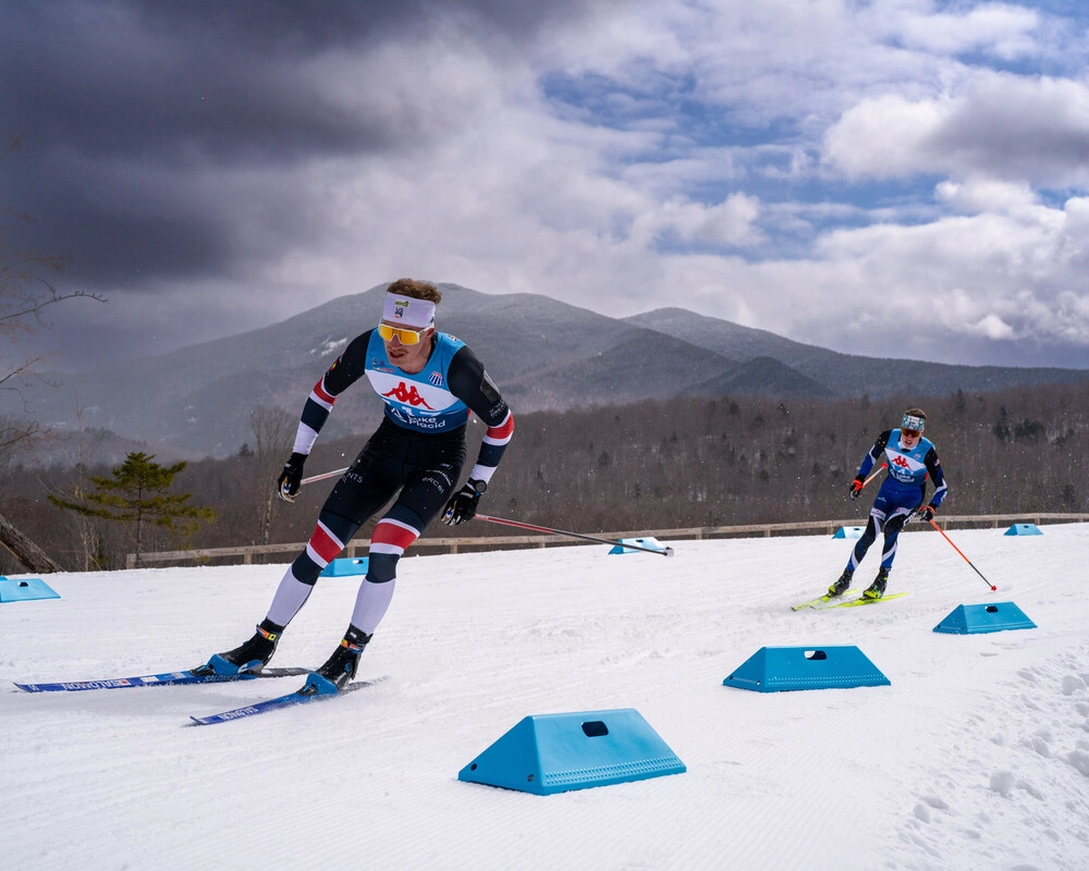 The image shows two cross-country skiers racing on a snowy course. 