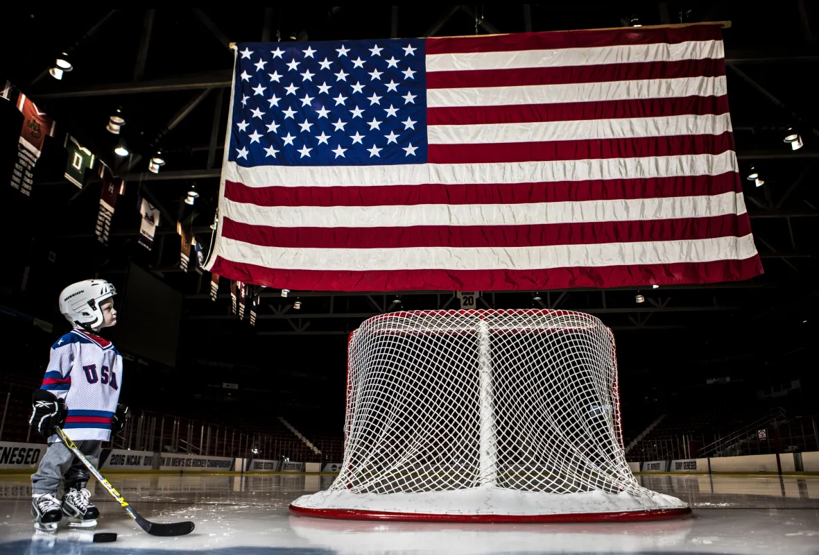 Young boy in hockey gear stands next to hockey goal faced away with large American flag flying above it in hockey rink on ice