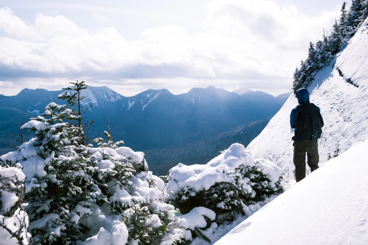 A man in full winter gear looks out from a snowy summit to a mountain range. 