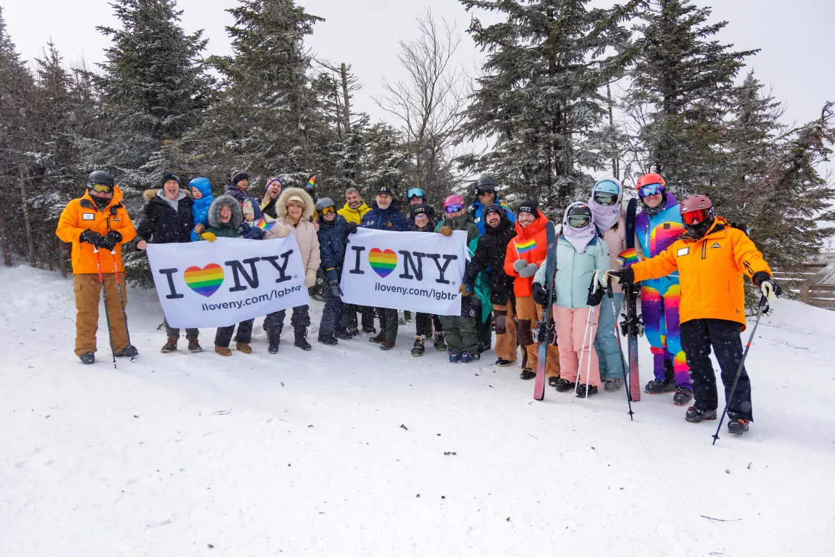 A group of adult skiers poses for a group photo on a snowy mountaintop, holding flags that read "I *heart* NY". The hearts are rainbow striped.