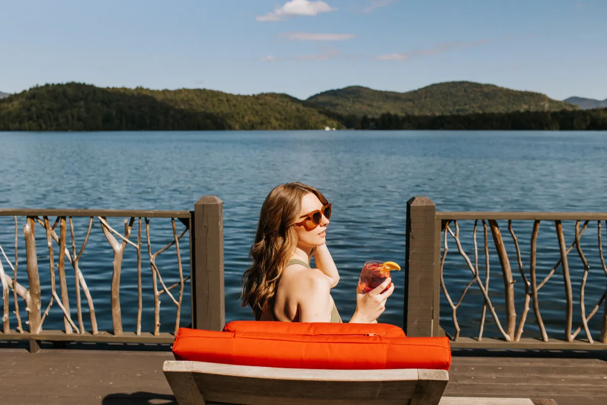 A woman sits on a cushy red chair on a rustic, twig-decorated deck overlooking a broad blue lake with low mountains in the distance.