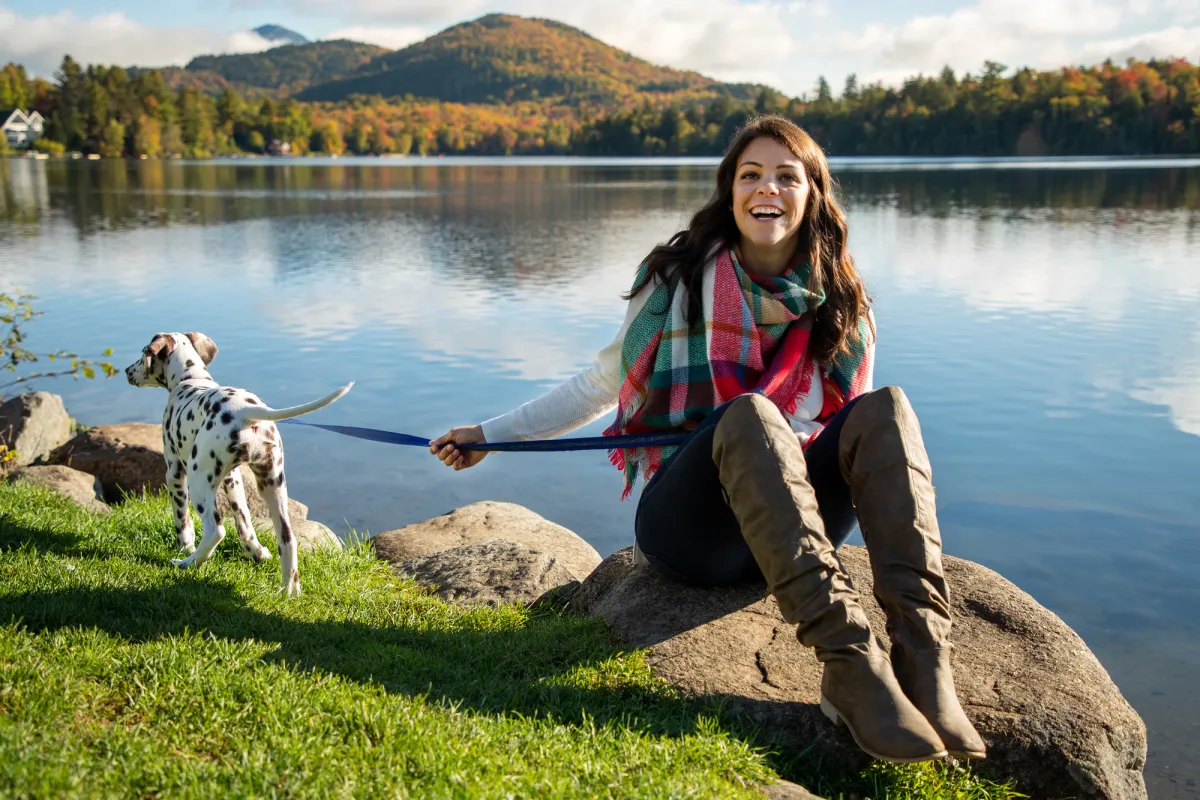 A woman in a fall outfit sits at the edge of a lake with her dalmation puppy on a leash in fall. 