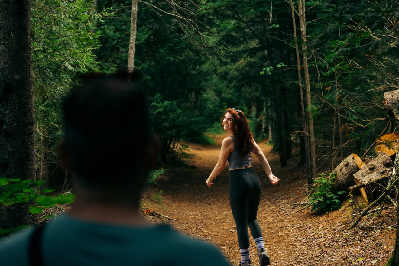 Young woman looks back over her shoulder at man on thickly forested trail in summer