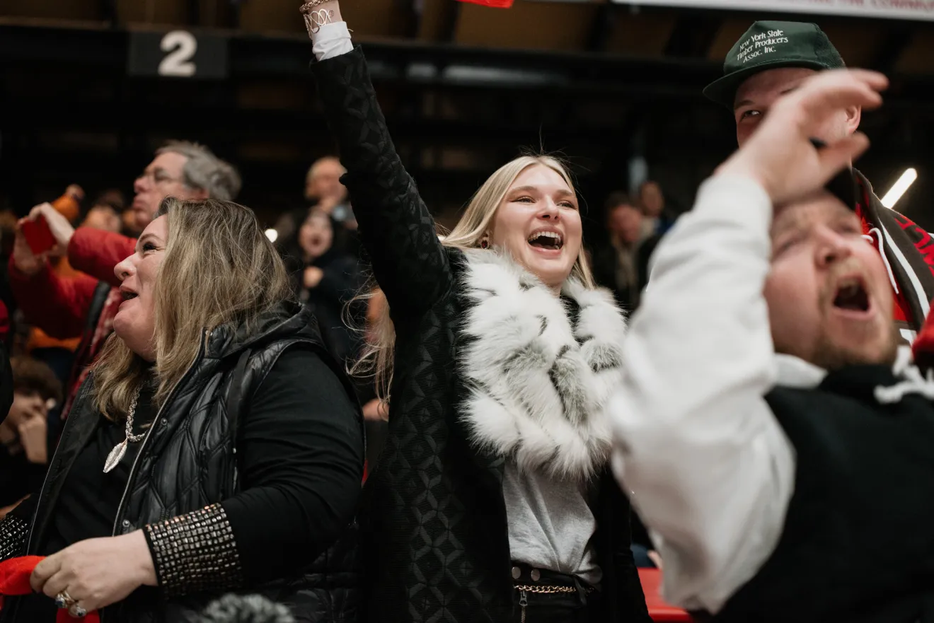 Young woman dressed in black coat and furry scarf cheers amidst packed crowd at hockey game