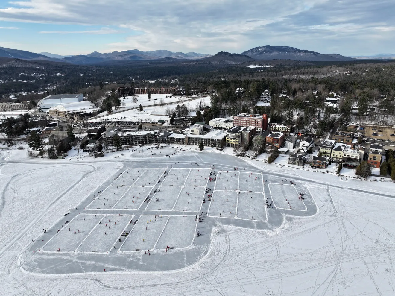 An aerial view of Mirror Lake frozen over and divided into numerous pond hockey rinks. A small town overlooks the lake.