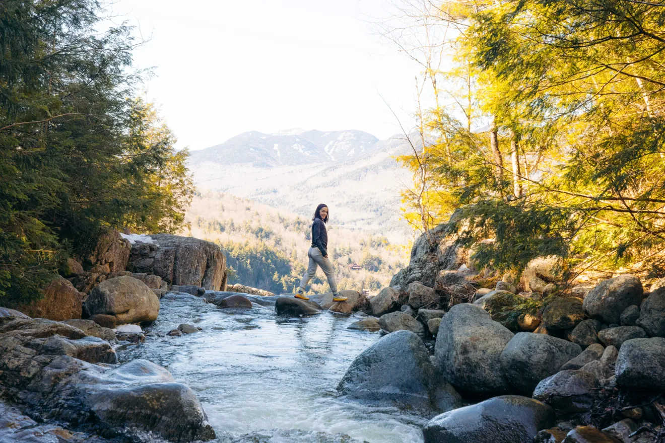 Young woman stands on rocks above stream at top of mountain waterfall with summits in the distance