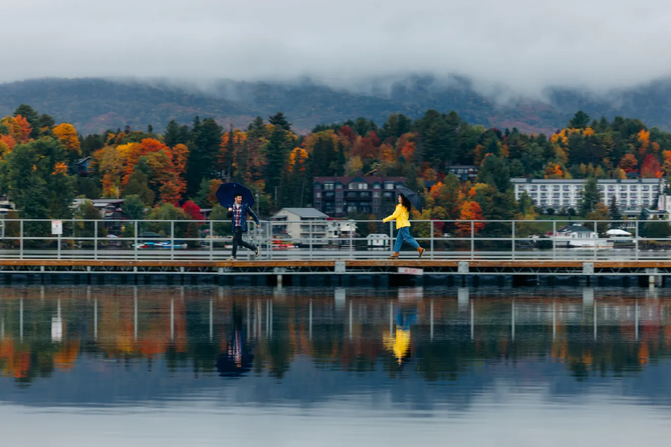 Man in black rain coat and woman in yellow rain coat, both with umbrellas, run along dock with Main Street and fall colors in background