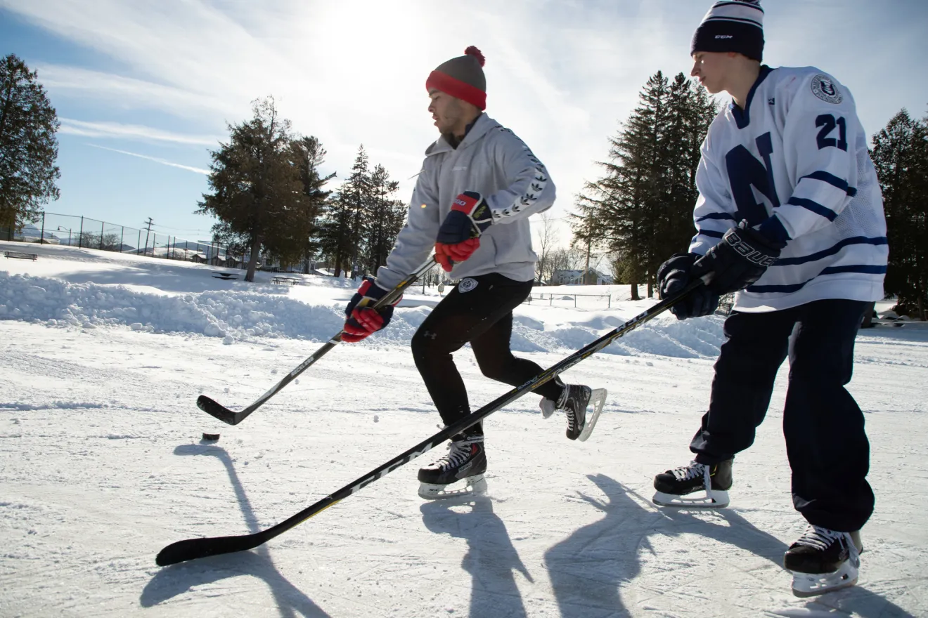 Two men play hockey on an icy pond. 