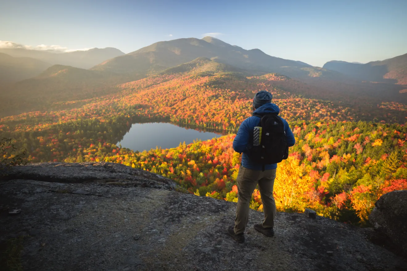 Hiker stands on summit of Mt Jo looking at view of Heart Lake and High Peaks with fall colors