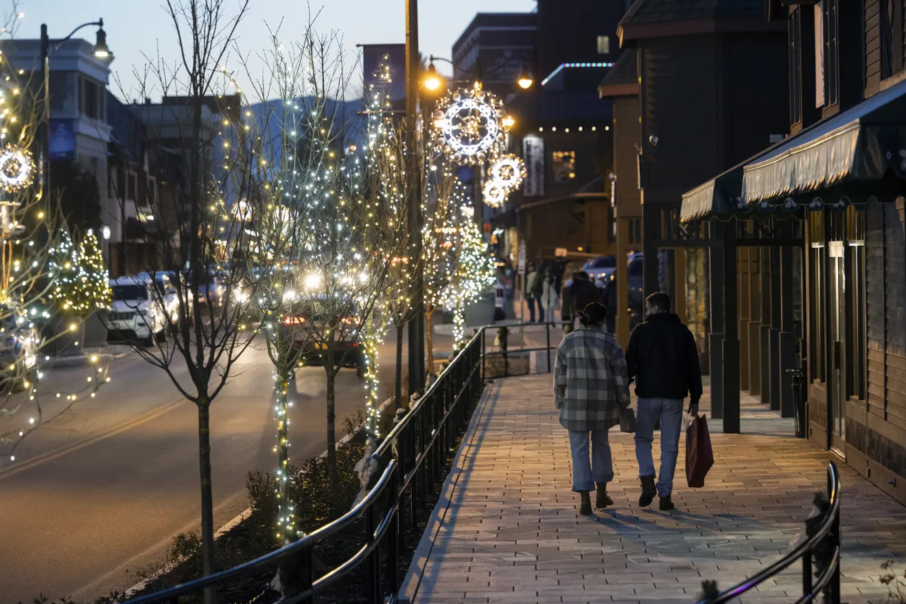 Lake Placid's Main Street is lit by festive snowflake lights on lampposts as shoppers stroll the sidewalk.