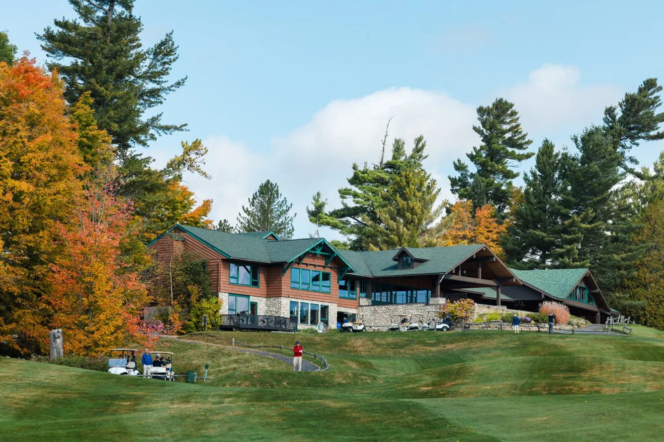 A wood and stone golf clubhouse is surrounded by green lawns and fall foliage.