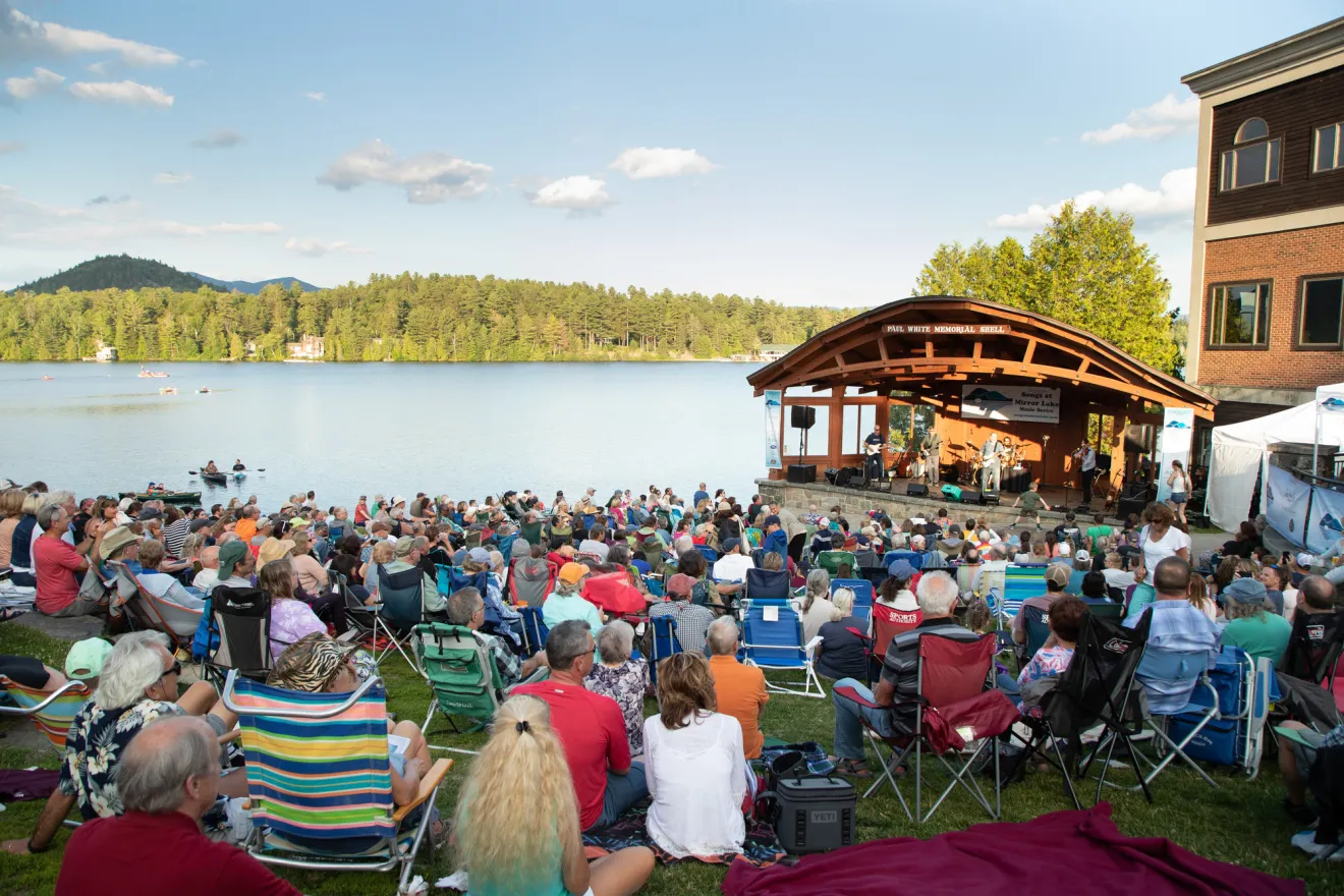 Crowd of people listening to music on lawn in front of bandshell next to lake with mountain views