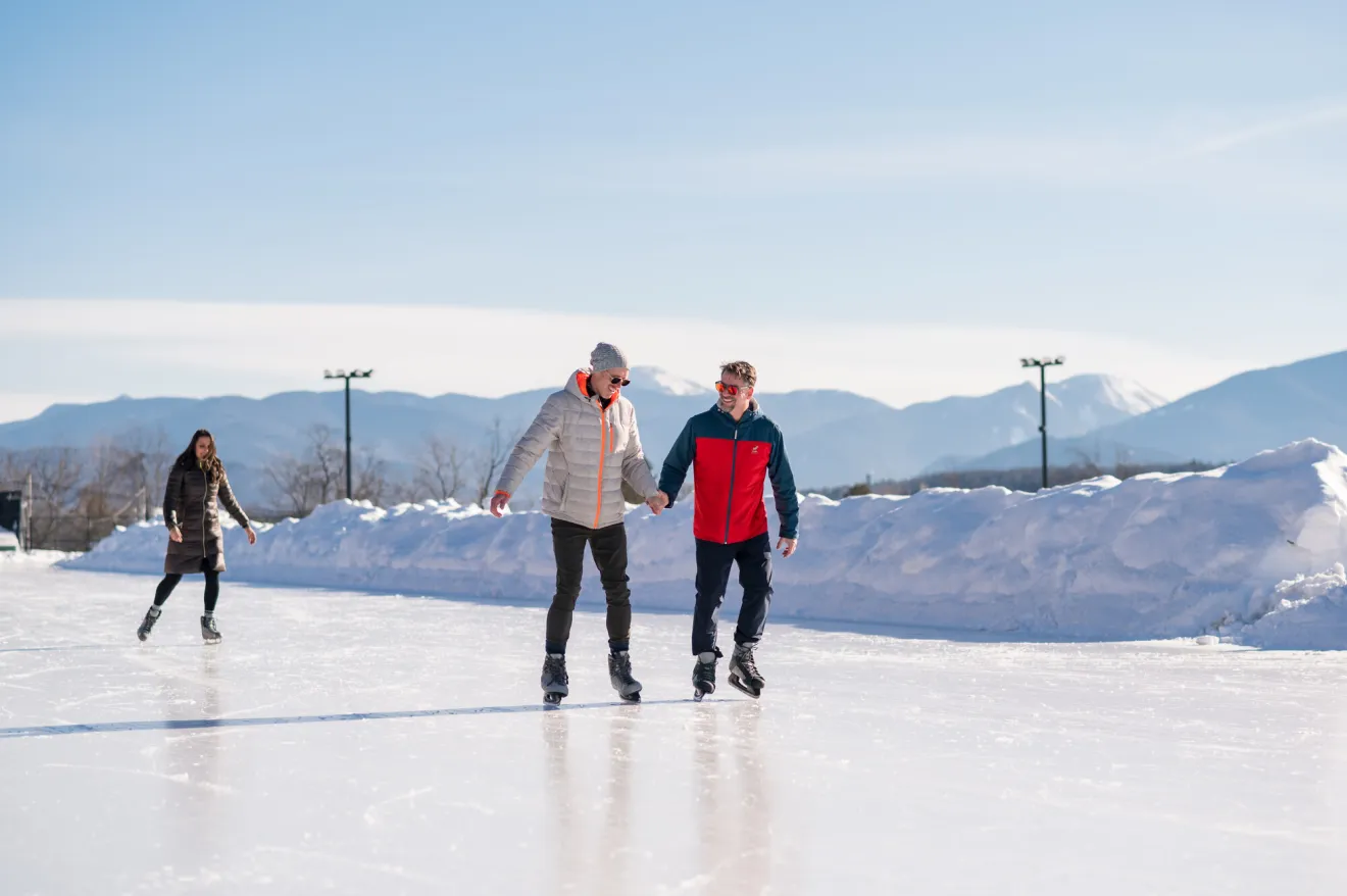 Two men hold hands while skating on an outdoor oval on a sunny winter day.
