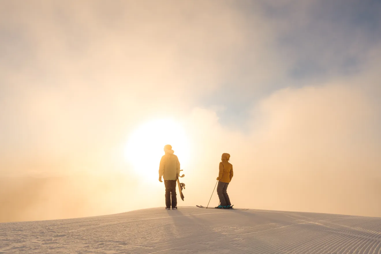 Skier and snowboarder stand at top of snowy mountain looking out at sunrise