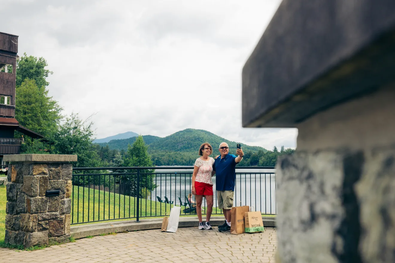 Senior couple, man and woman, stand at park railing overlooking lake and mountains taking a selfie with shopping bags