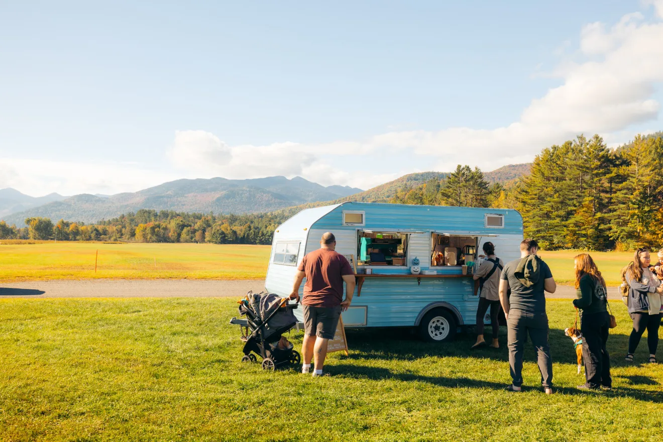 A retro camper is parked on a field on a sunny summer day. People casually line up to purchase fresh produce from the camper.