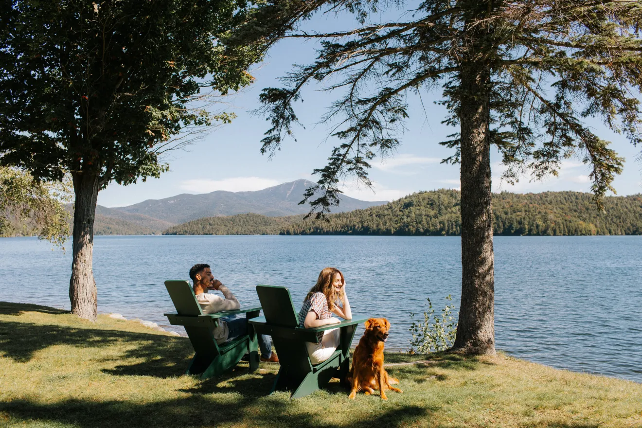 Couple sits next to lake in green wooden chairs under pine trees with golden retriever dog