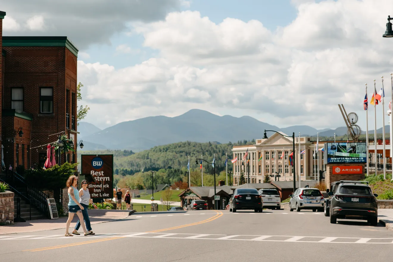 Two people walking across the street in Lake Placid, with the mountains in the background.