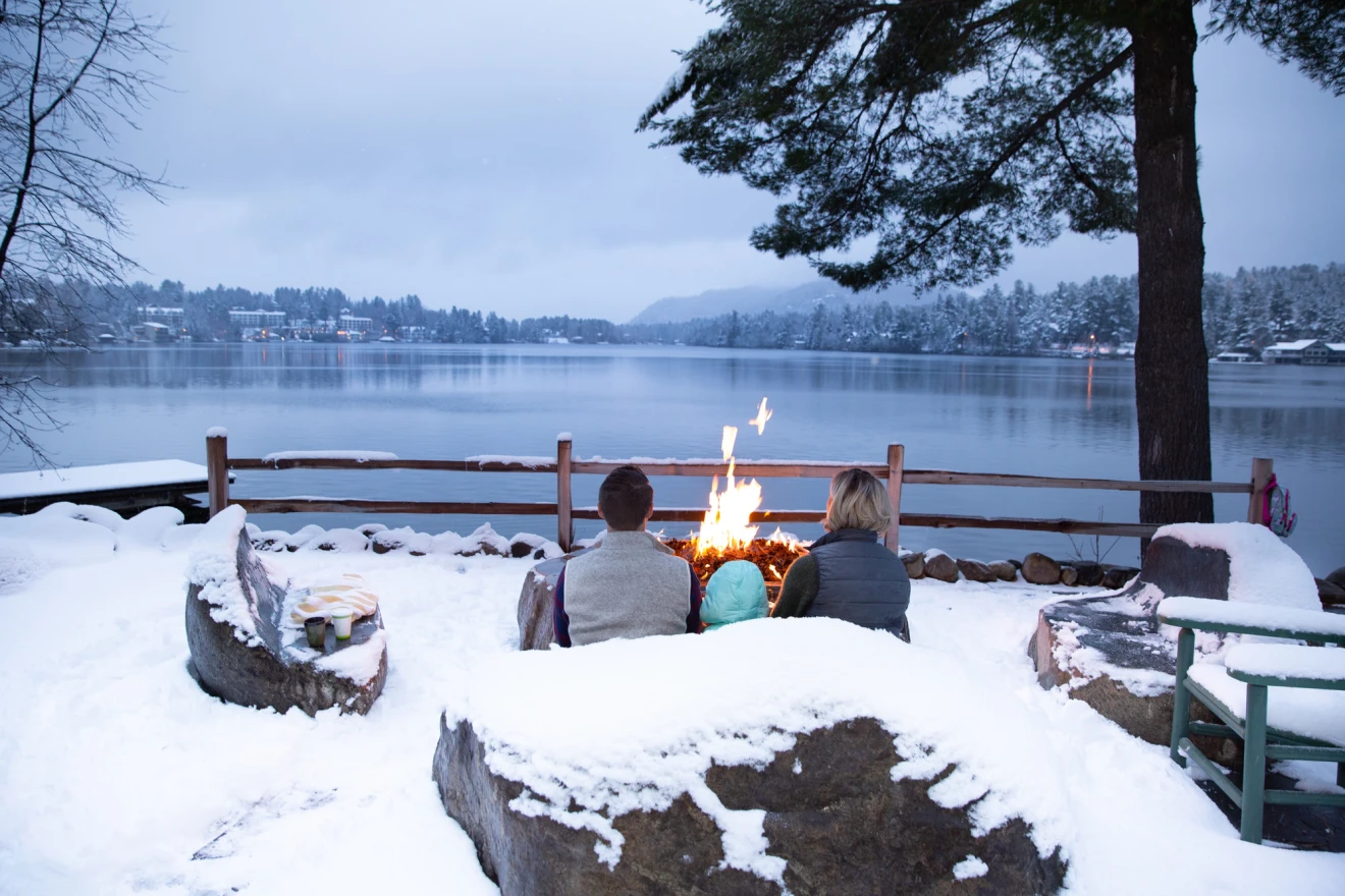 A couple sitting in front of a fire, looking out at Mirror Lake during their stay in Lake Placid.