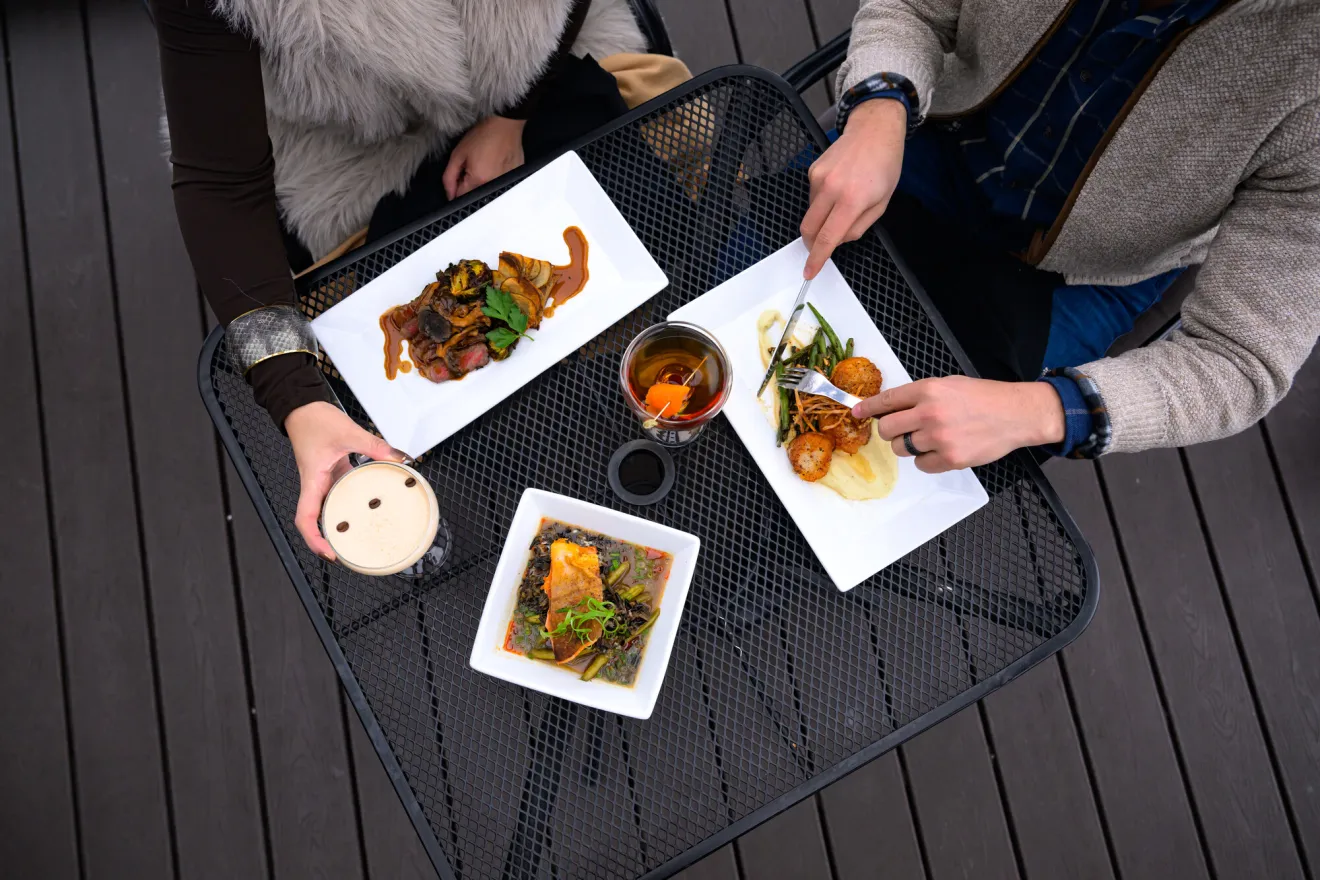 Top down view of 3 small plates of food and cocktails on a black wire table as two people eat and drink next to each other