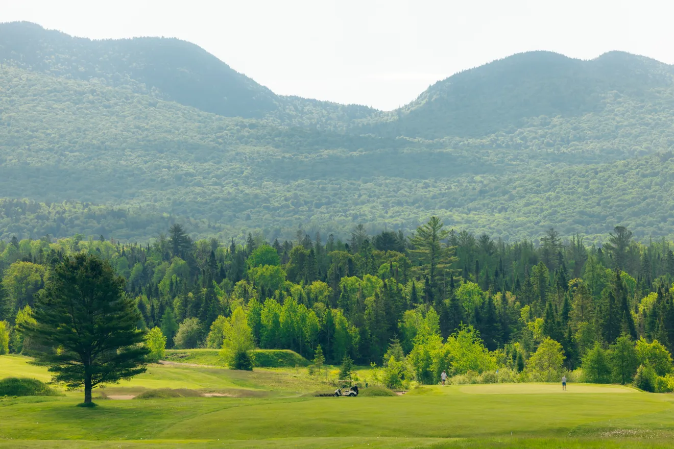 A wide view of a lush golf course in May, with high, forested mountains in the background.