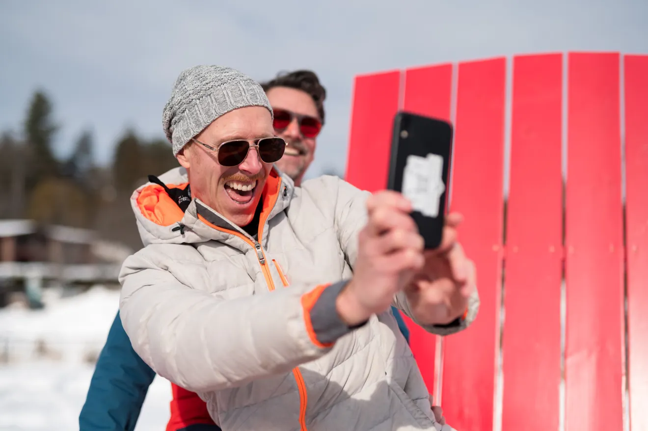 Two men stand on giant red Adirondack chain in snow gear taking a self on a cell phone