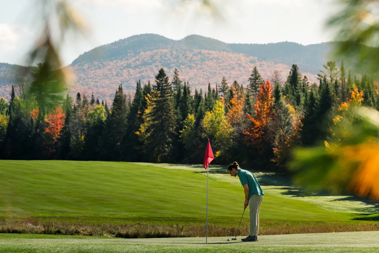 A golfer prepares to putt on a bright green course with a colorful, fall foliage-covered mountain in the background.