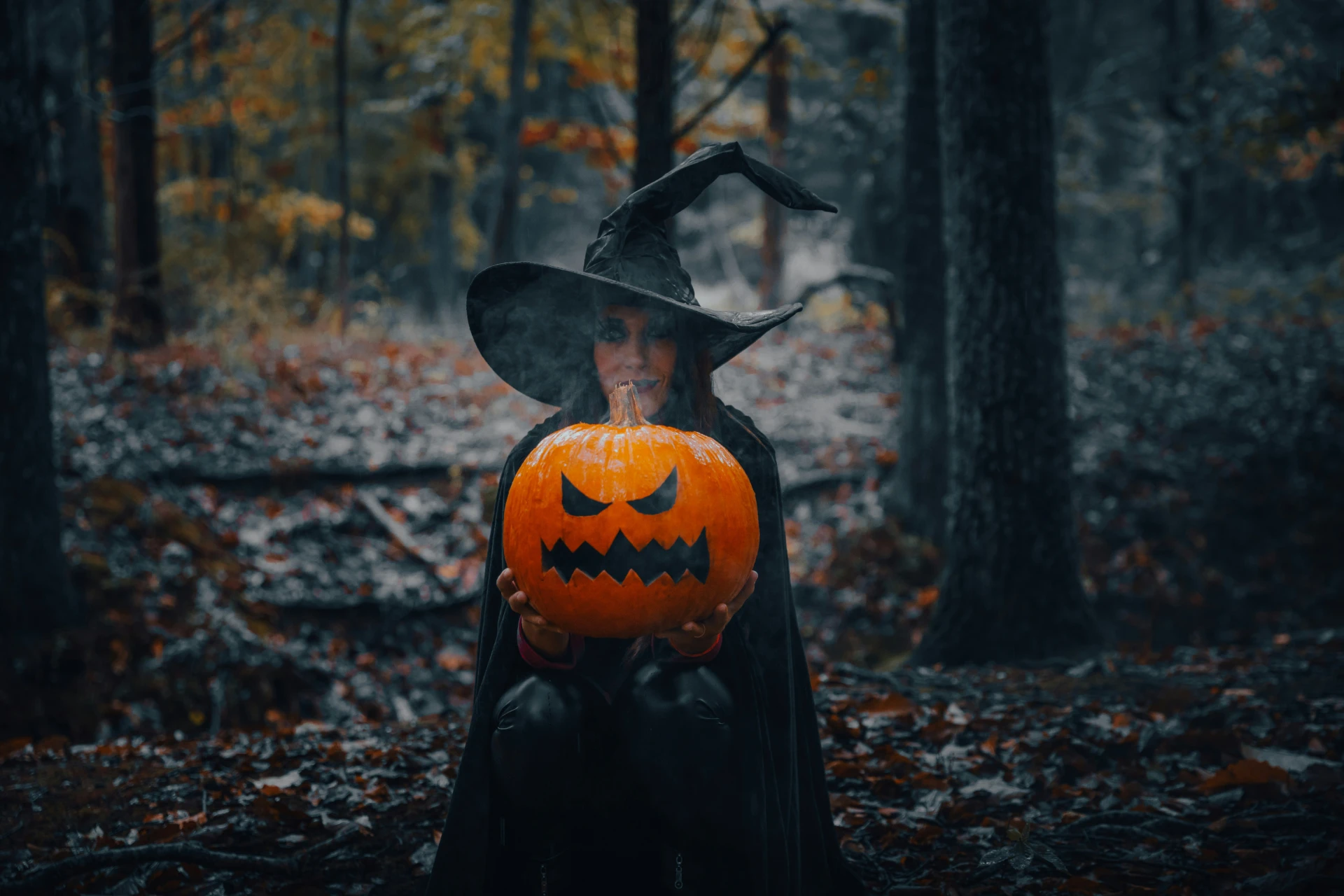Woman stands in woods in black witch robes and hat holding large jack-o-lantern