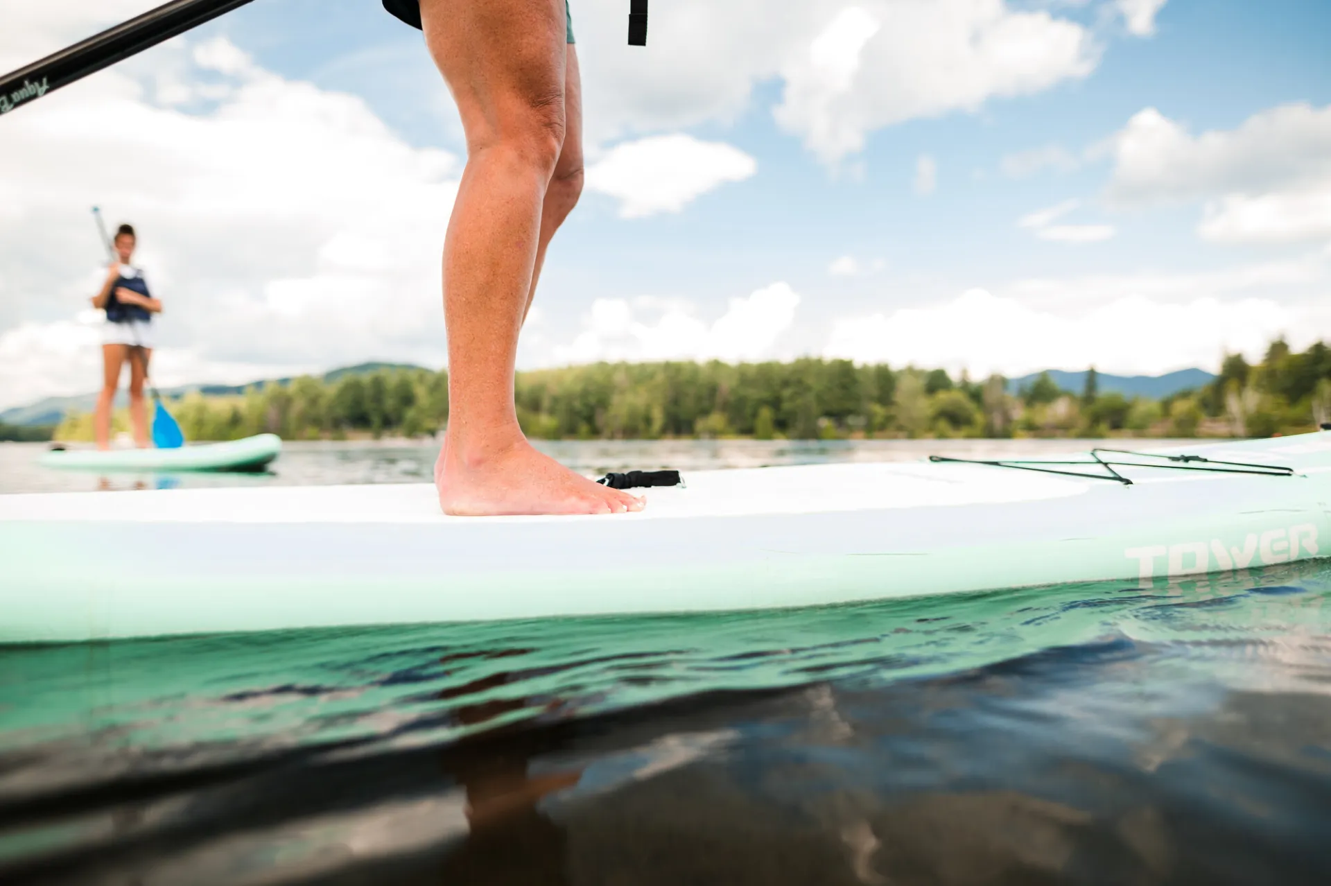 Person's feet and legs seen standing on a white paddle board with a second person paddling a paddle board in the background