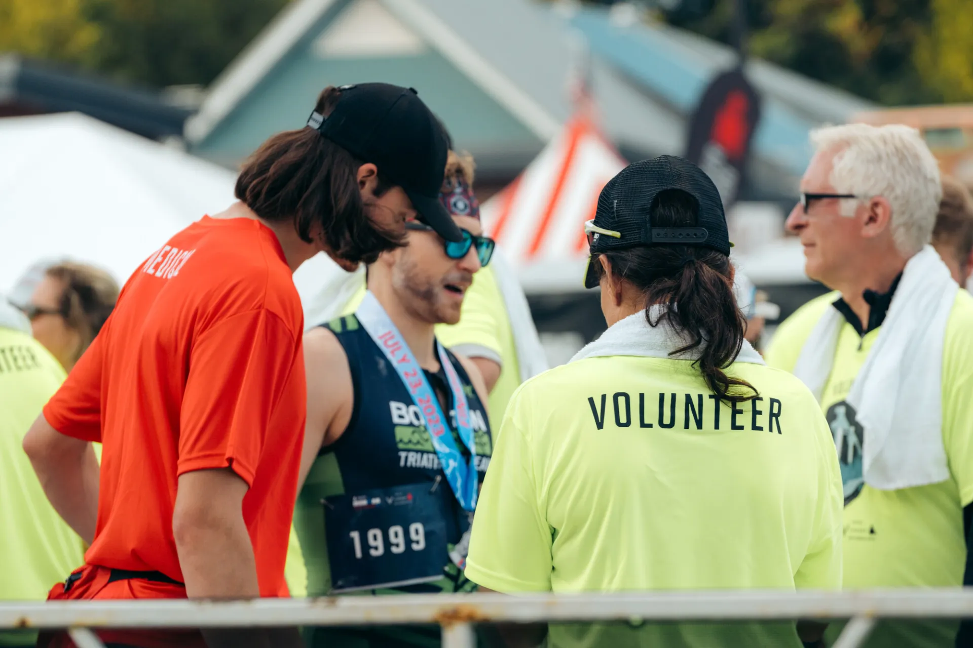 Ironman volunteers wearing bright yellow shirts with the word volunteer across the shoulders seen from behind talking to finisher wearing medal
